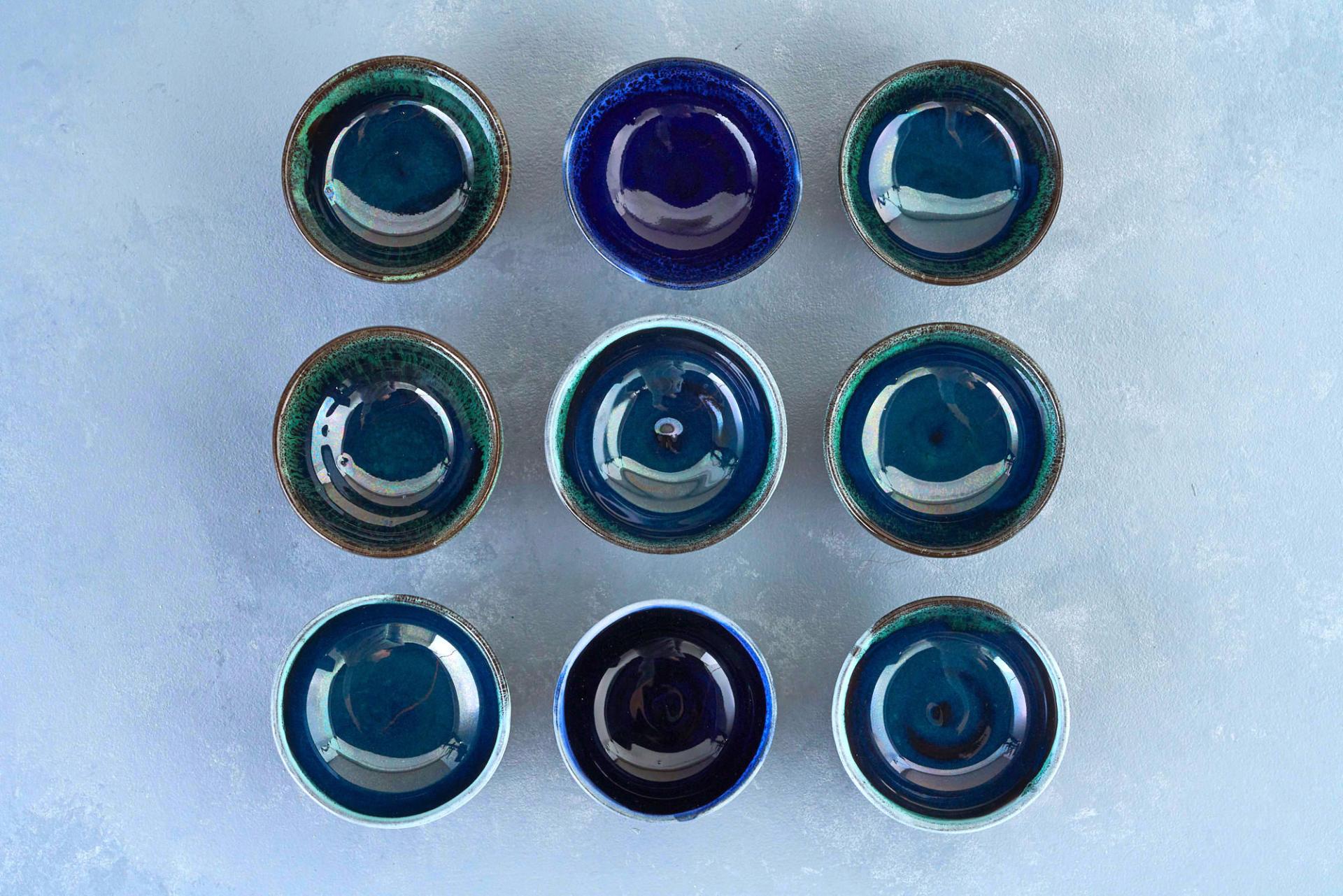 Nine handcrafted ceramic bowls in various shades of blue arranged in a grid on a textured surface.