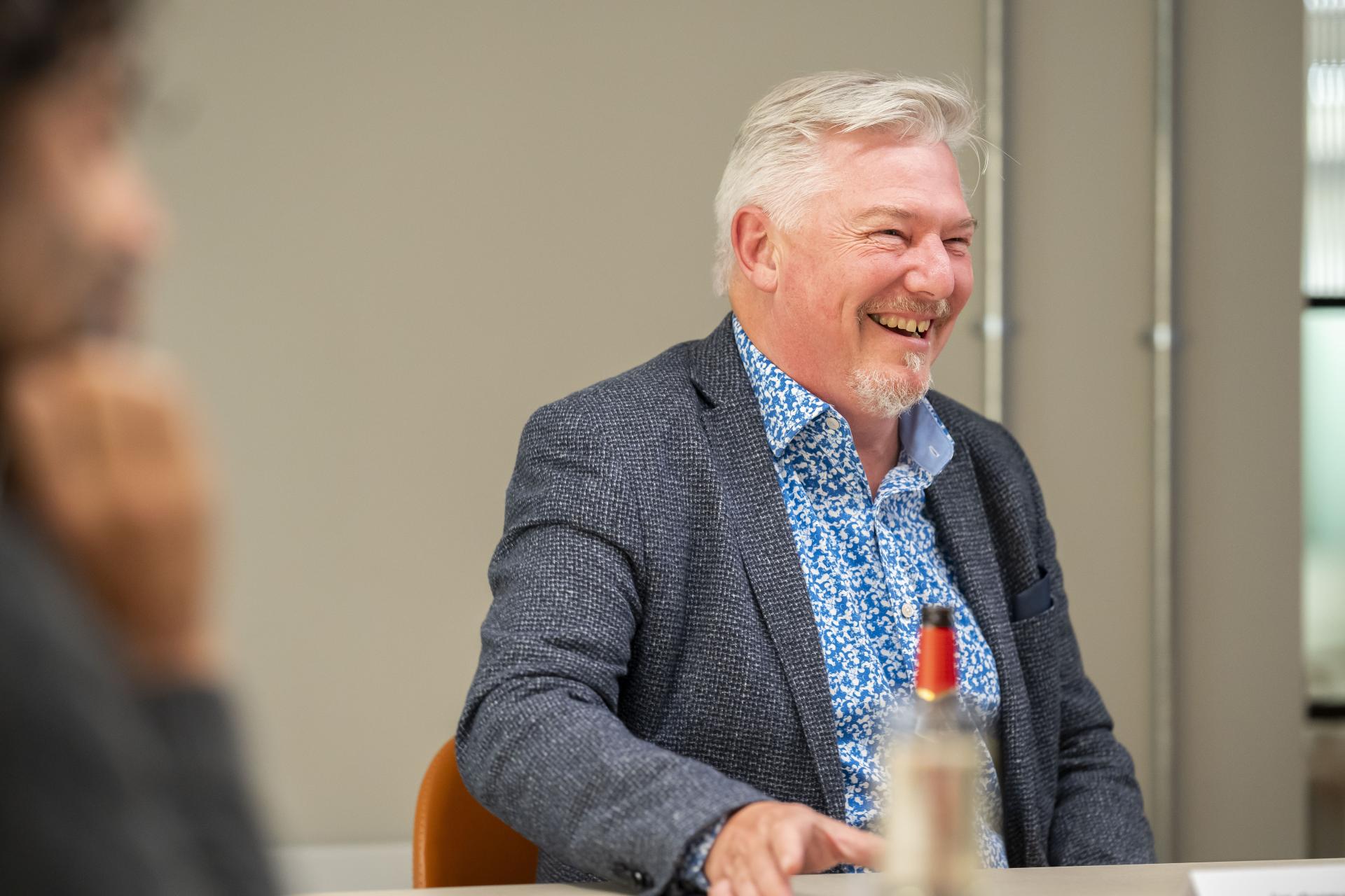 Smiling man in a blue patterned shirt discusses workplace impact of data at a business meeting table.