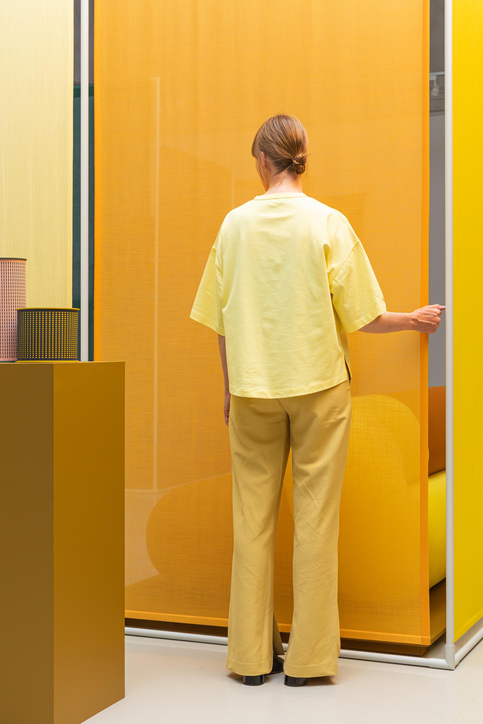 A woman in yellow attire interacts with vibrant bio-based materials in an innovative design space, reflecting on color palettes.