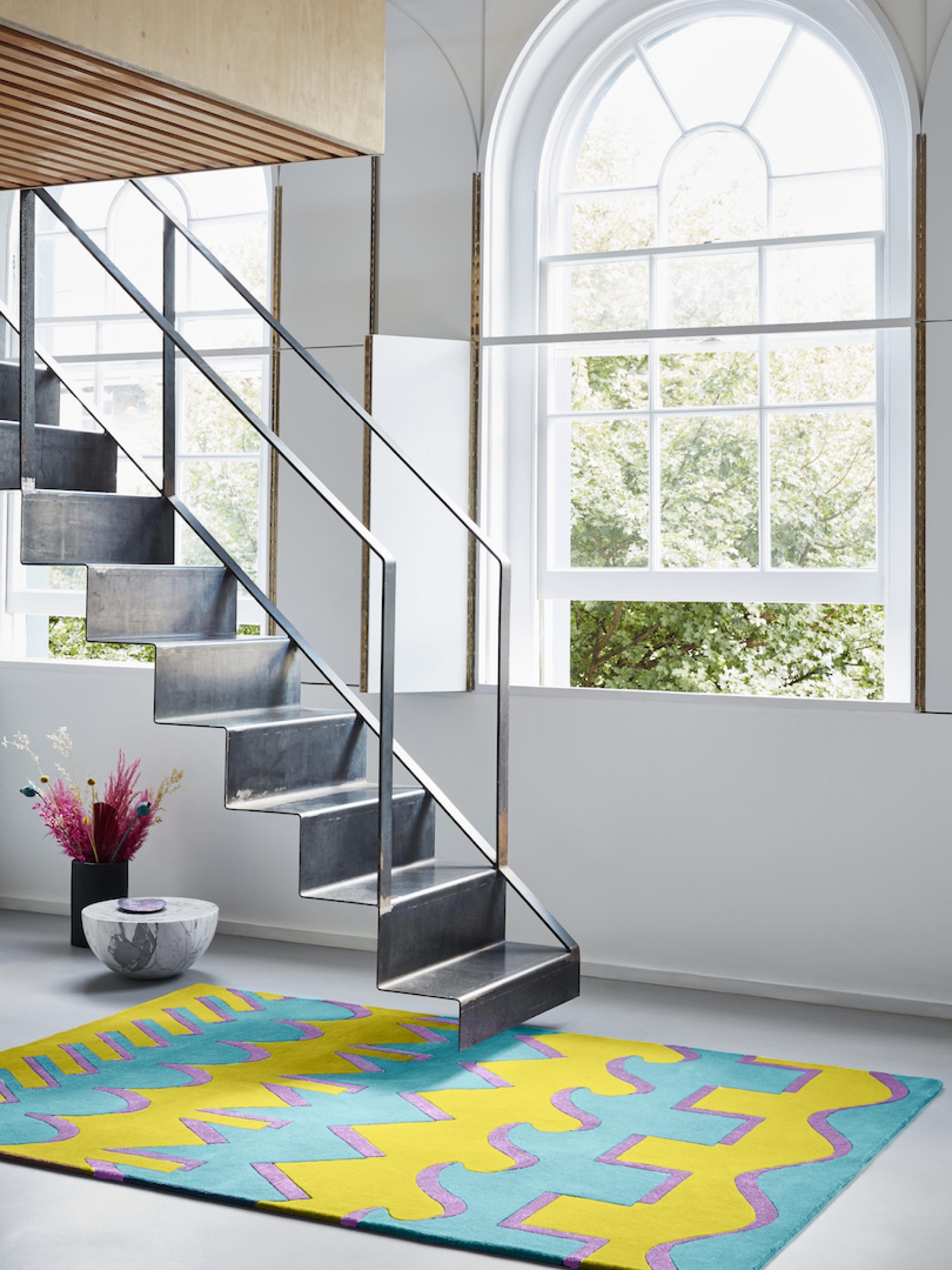 Vibrant Mediterranean-inspired rug by Adam Nathaniel Furman, showcased under a modern staircase with large windows.