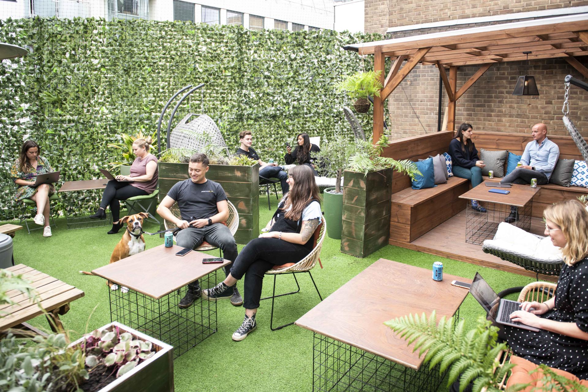 A vibrant outdoor workspace featuring diverse individuals collaborating, a dog, and lush greenery, highlighting modern work environments.