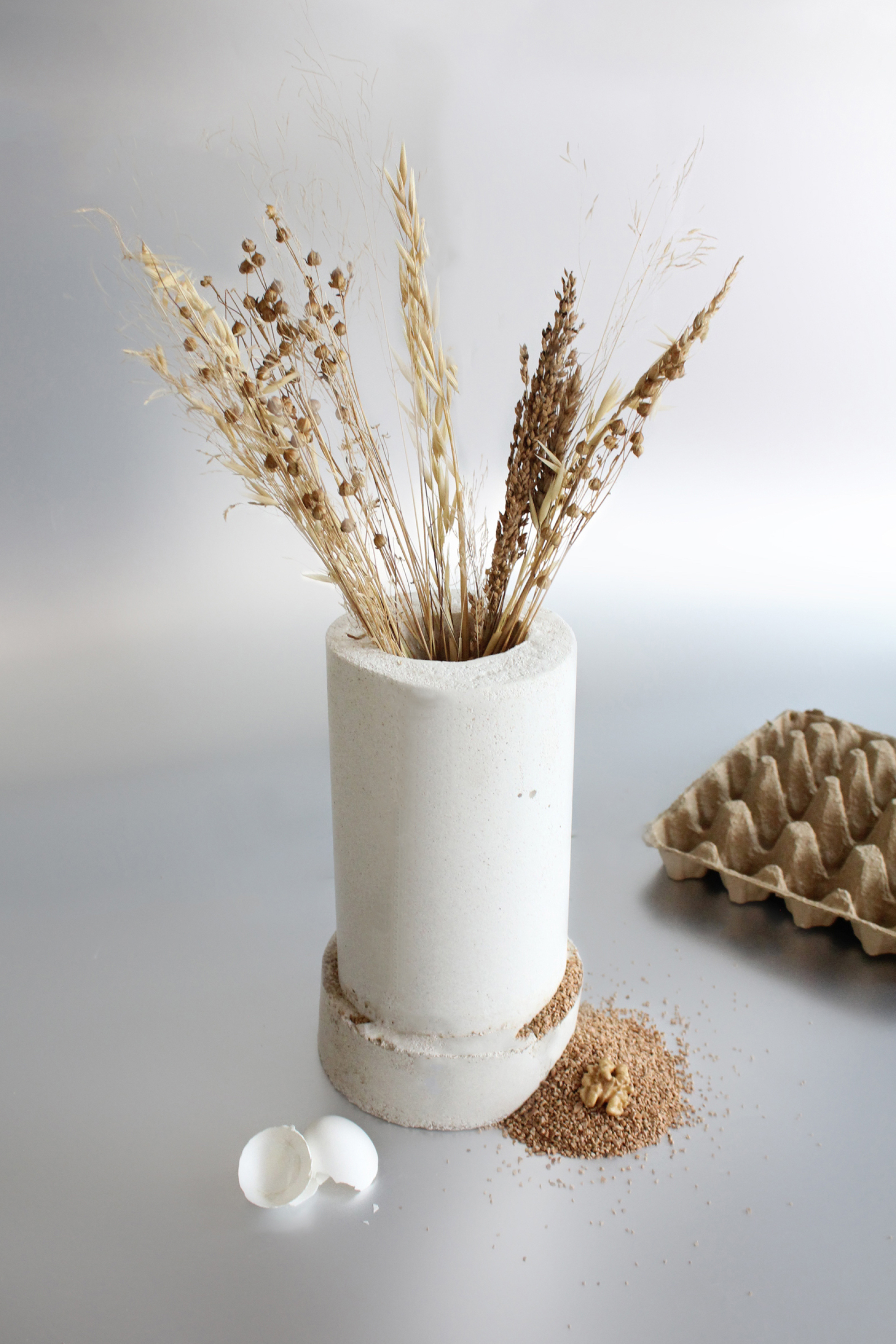 A minimalist vase made from bio-based materials, holding dried plants, with egg shells and compost nearby.