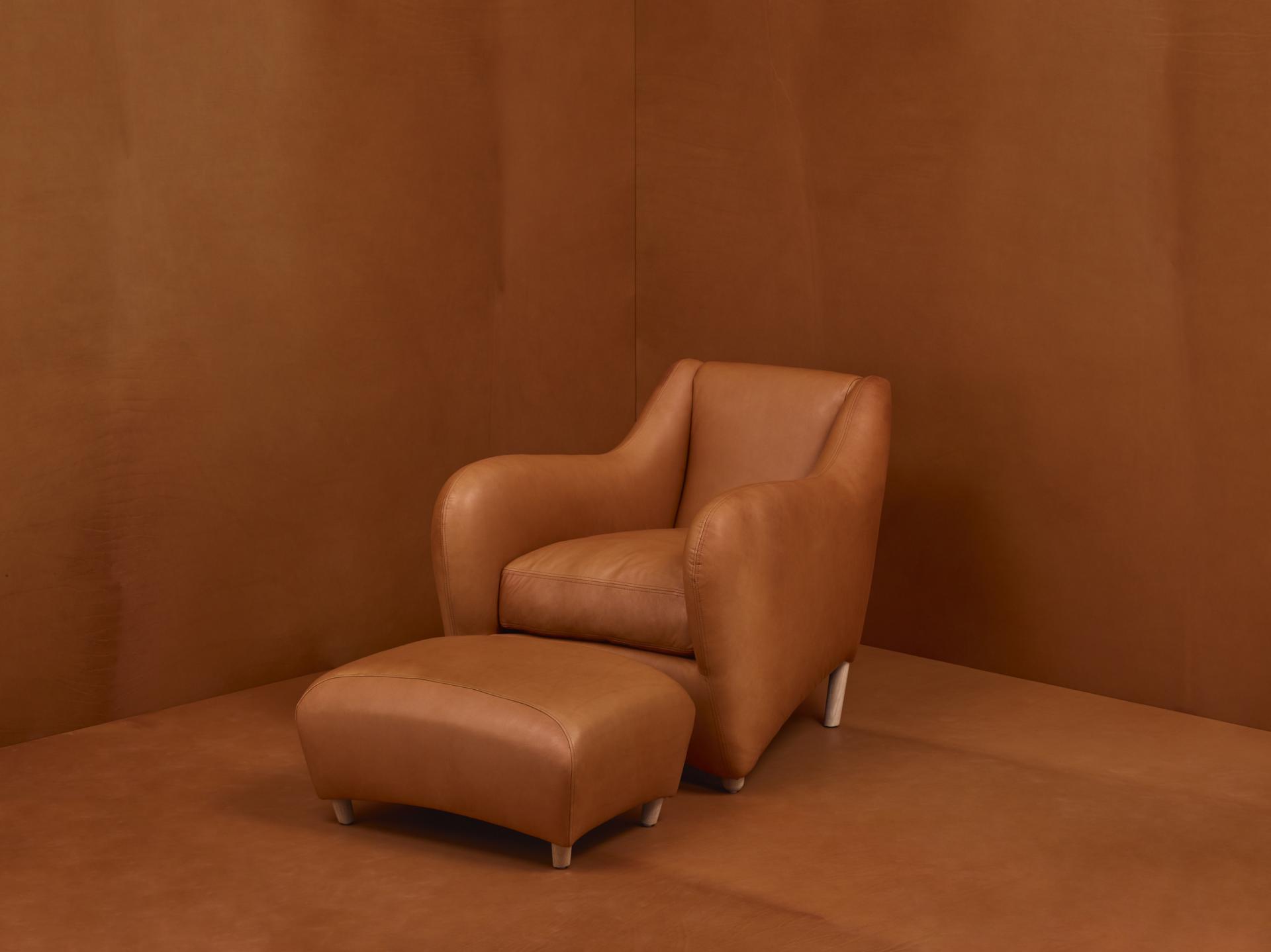 Matthew Hilton's Balzac armchair in warm leather with matching ottoman against a textured brown backdrop.