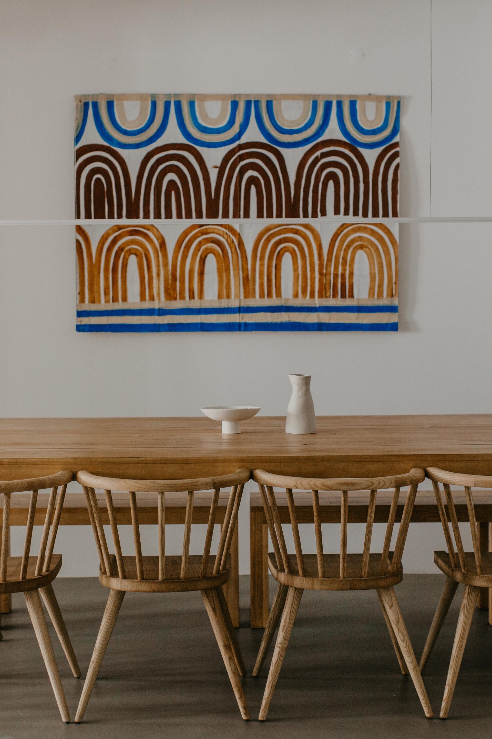Wooden dining table with minimalist decor, featuring ceramic vase and bowl, complemented by a colorful abstract art piece.