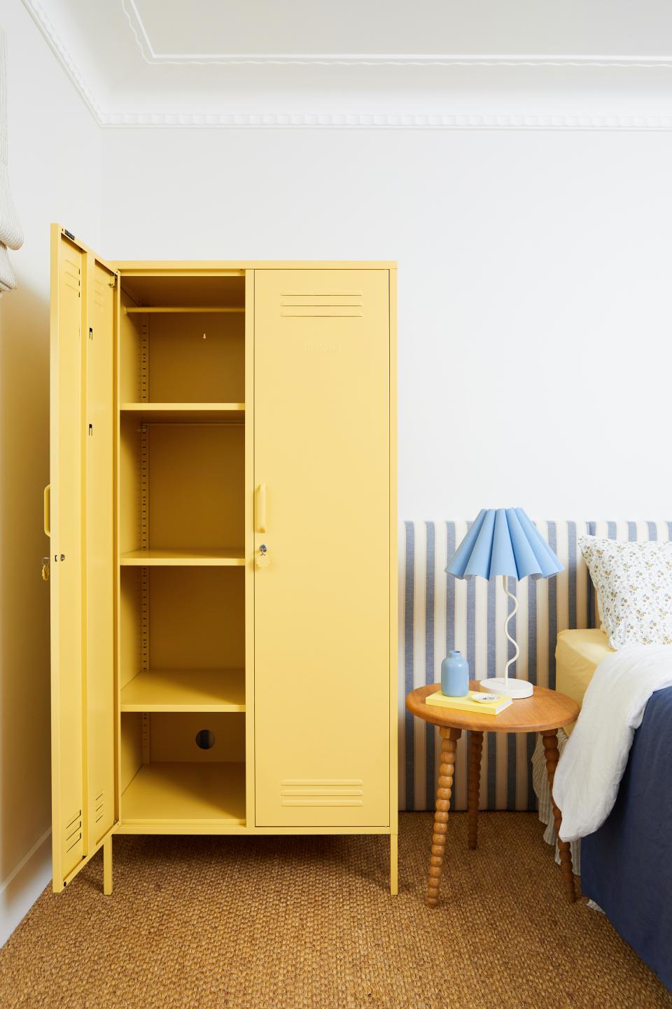 Mustard Made's new 'Butter' yellow locker stands open beside a stylish bedside table and blue lamp in a cozy room.