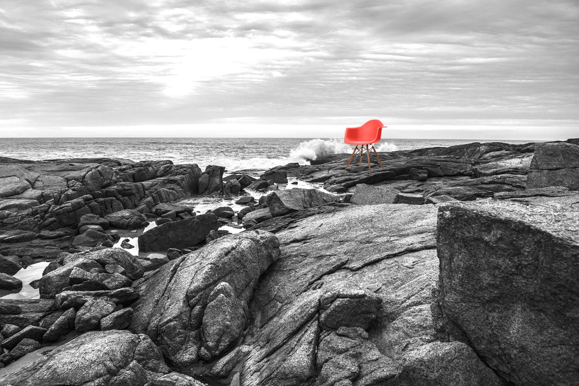 Bright red design chair placed on rocky shore against a dramatic ocean backdrop, highlighting innovative workspace aesthetics.