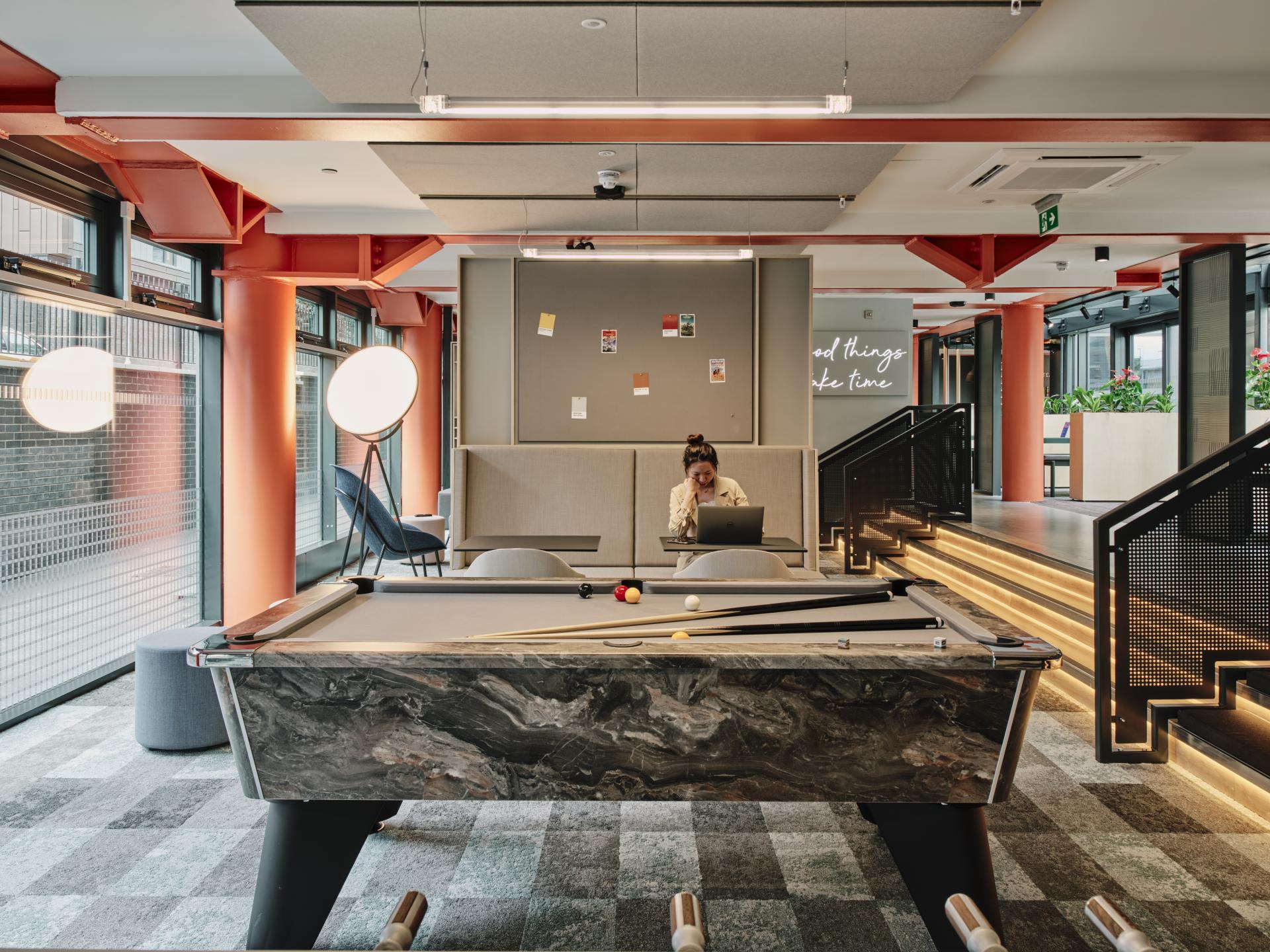 Modern workspace with a stylish pool table, featuring a woman working on a laptop in a creative environment.