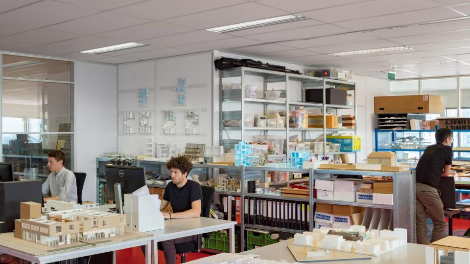 Modern architectural studio workspace featuring models, sketches, and architects collaborating on innovative design projects.
