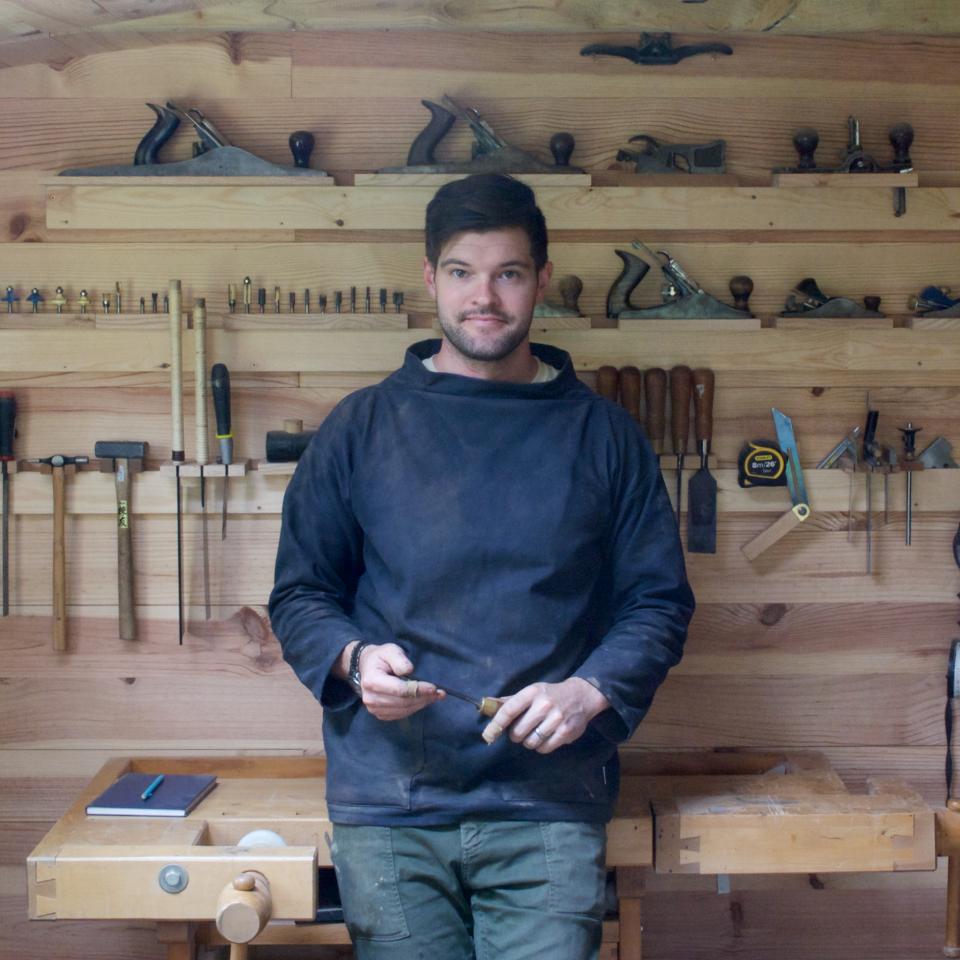 Contemporary furniture maker Sand Buchanan standing in his workshop, surrounded by woodworking tools and materials for sustainable design.
